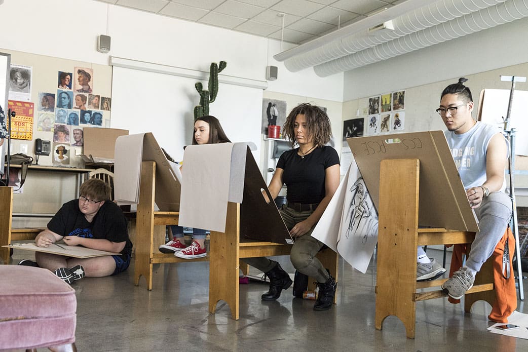 A row of students sitting in a classroom painting on easels