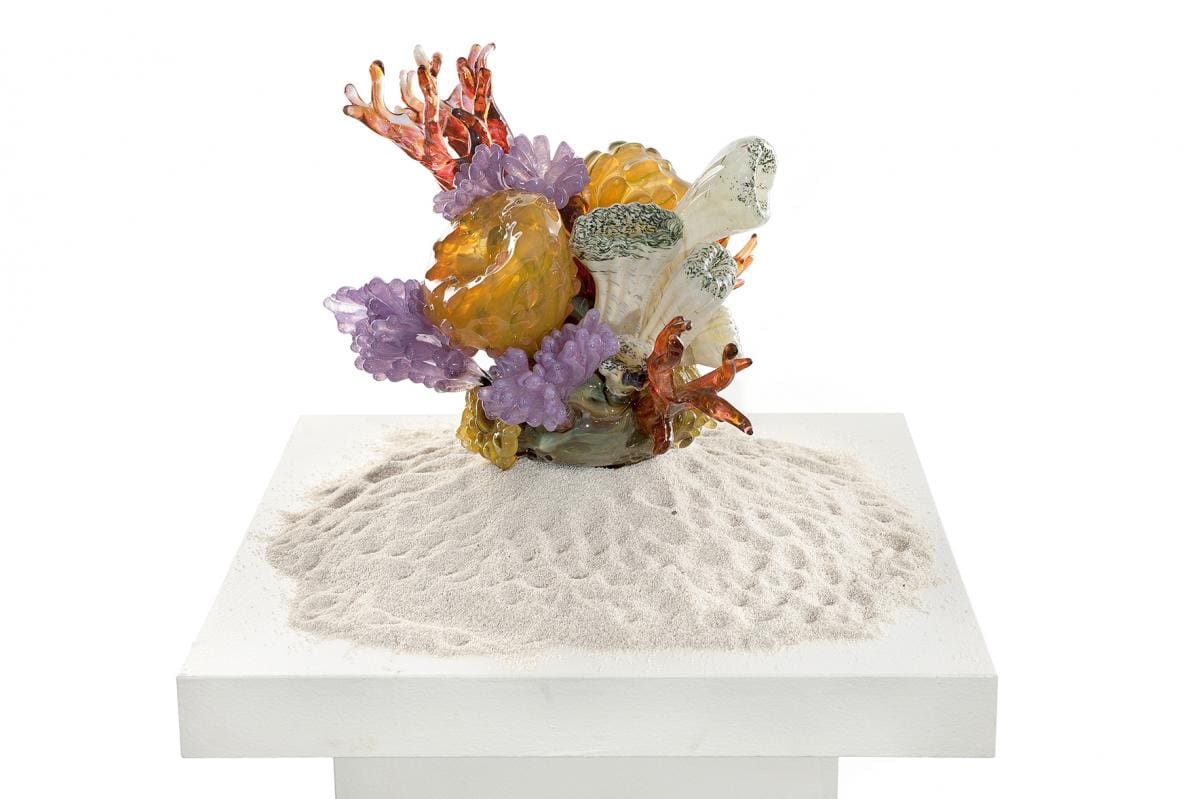 Glass sculpture on a pile of white sand on a white display table. The sculpture is made up of multiple yellow, purple, white and red pieces resembling coral in a coral reef.
