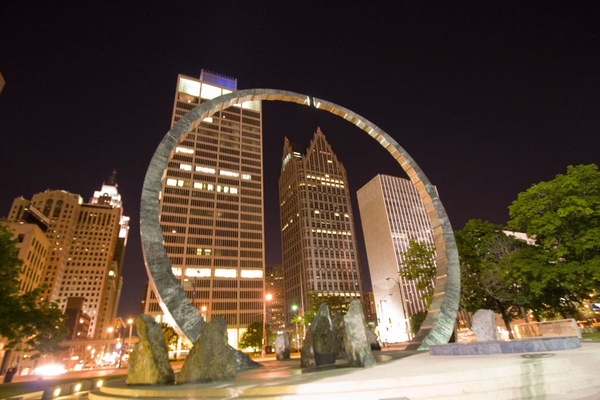 Photo of a huge metal ring sculpture in Detroit at night. It is pictured in front of a group of high rise building in the downtown.
