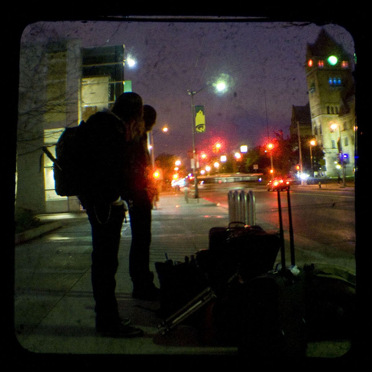 Photo of two people under an overpass in the middle of the night. They are looking down at a pile of luggage. The scene is lit up by streetlights.