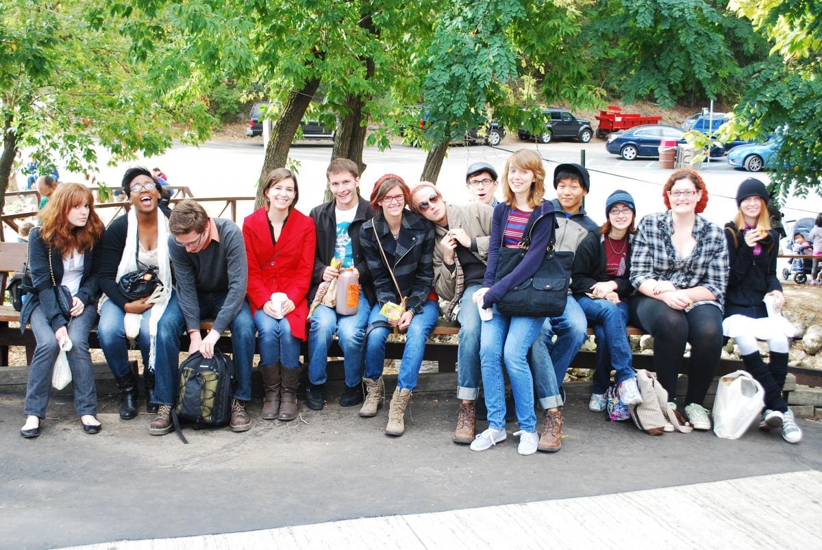 Photo of a large group of students sitting on a bench together and smiling for a picture. They are in a park parking lot.