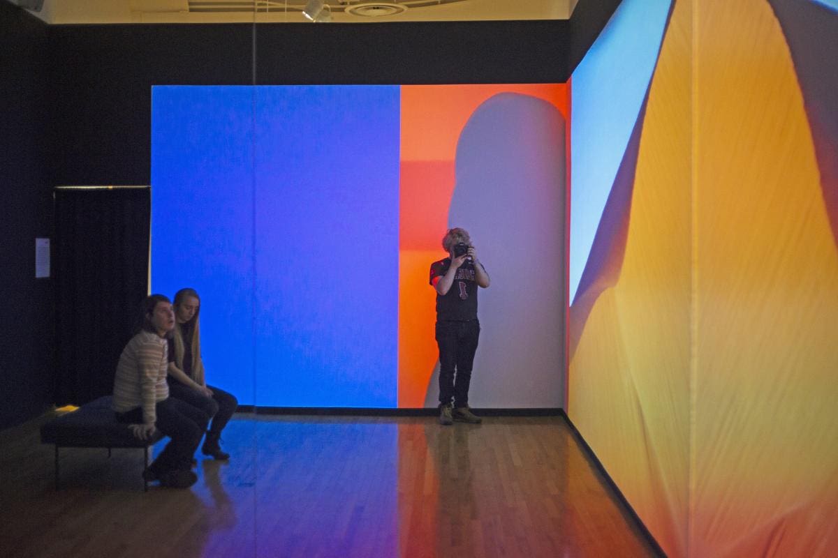 Two people on a bench and a person taking a photo in an art exhibition. They are admiring a large orange and blue projection on the wall.