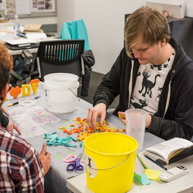 Two people sit at a table in a classroom and create a project using small blue, yellow and orange folded pieces of paper.