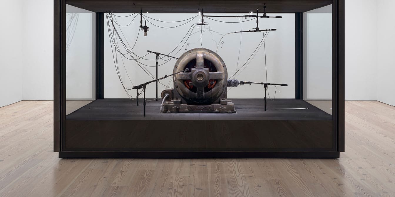 Large metal sculpture with countless wires coming out of the top inside of a box with glass panels and black metal frame. Pictured in an otherwise empty exhibition room.
