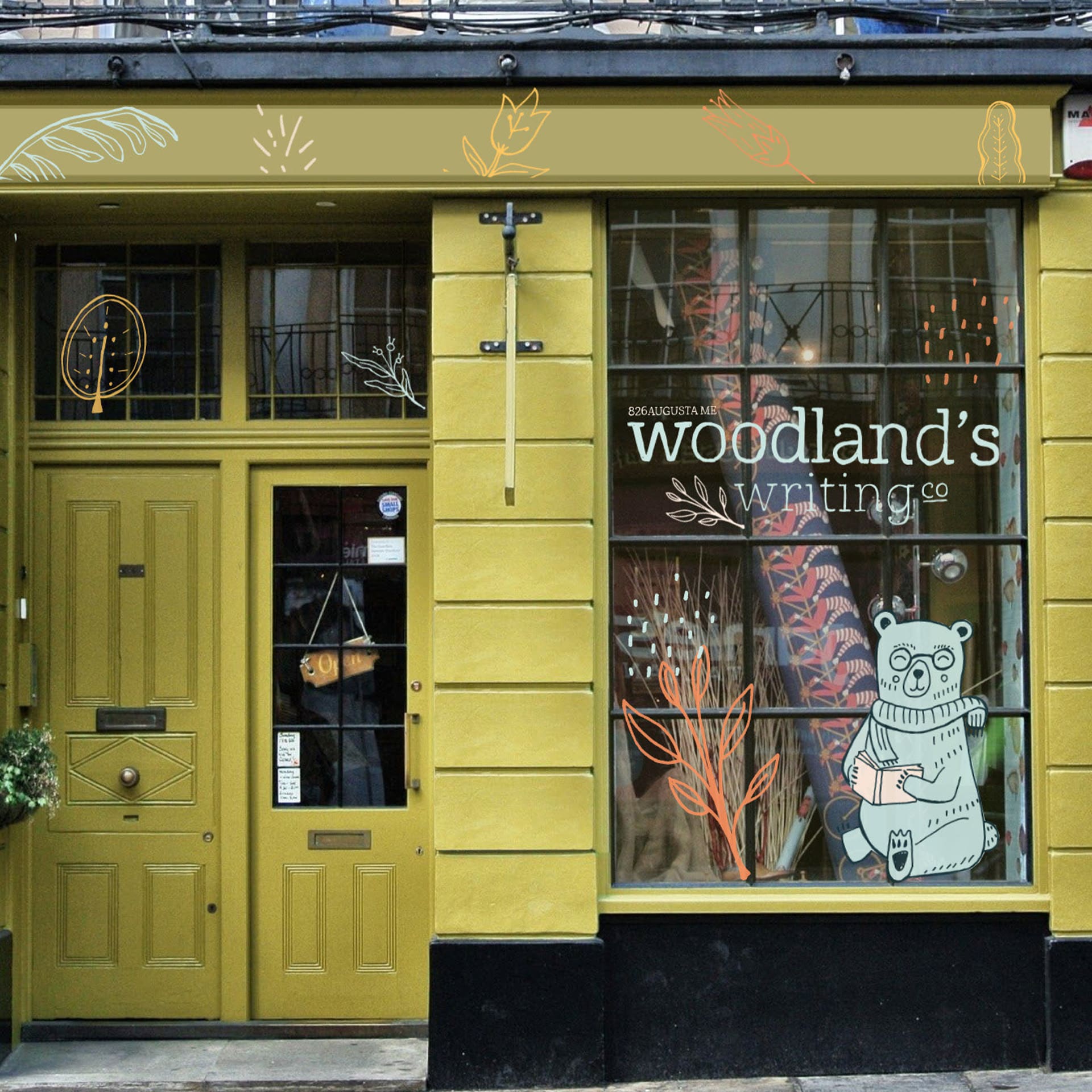 A yellow store front with a blue cartoon bear and orange branch sticker on the display window. The bear is sitting, smiling, reading a book, and wearing a scarf and glasses. There is also a sticker that reads the store's name, "Woodland's Writing Co." in lowercase letters.