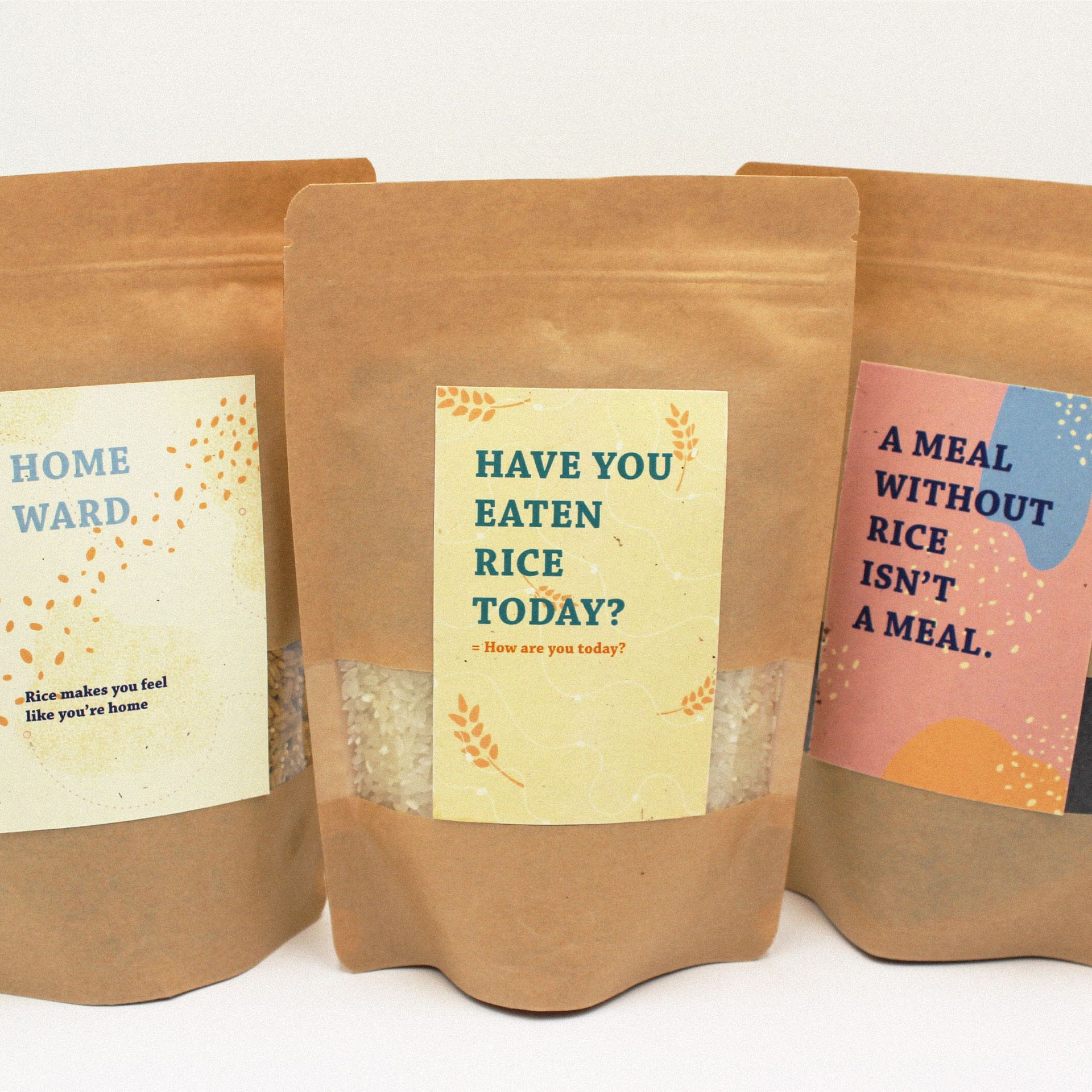 Photo of three brown paper bag packages of rice against a white background. each have a different rectangular label- the left is off-white, the center is yellow, and the right is pink. Each has different abstract shape decorations with a rice grain motif. The center bag reads "Have you eaten rice today?" in large blue text. The right bag reads 'A meal without rice isn't a meal." in large black text.