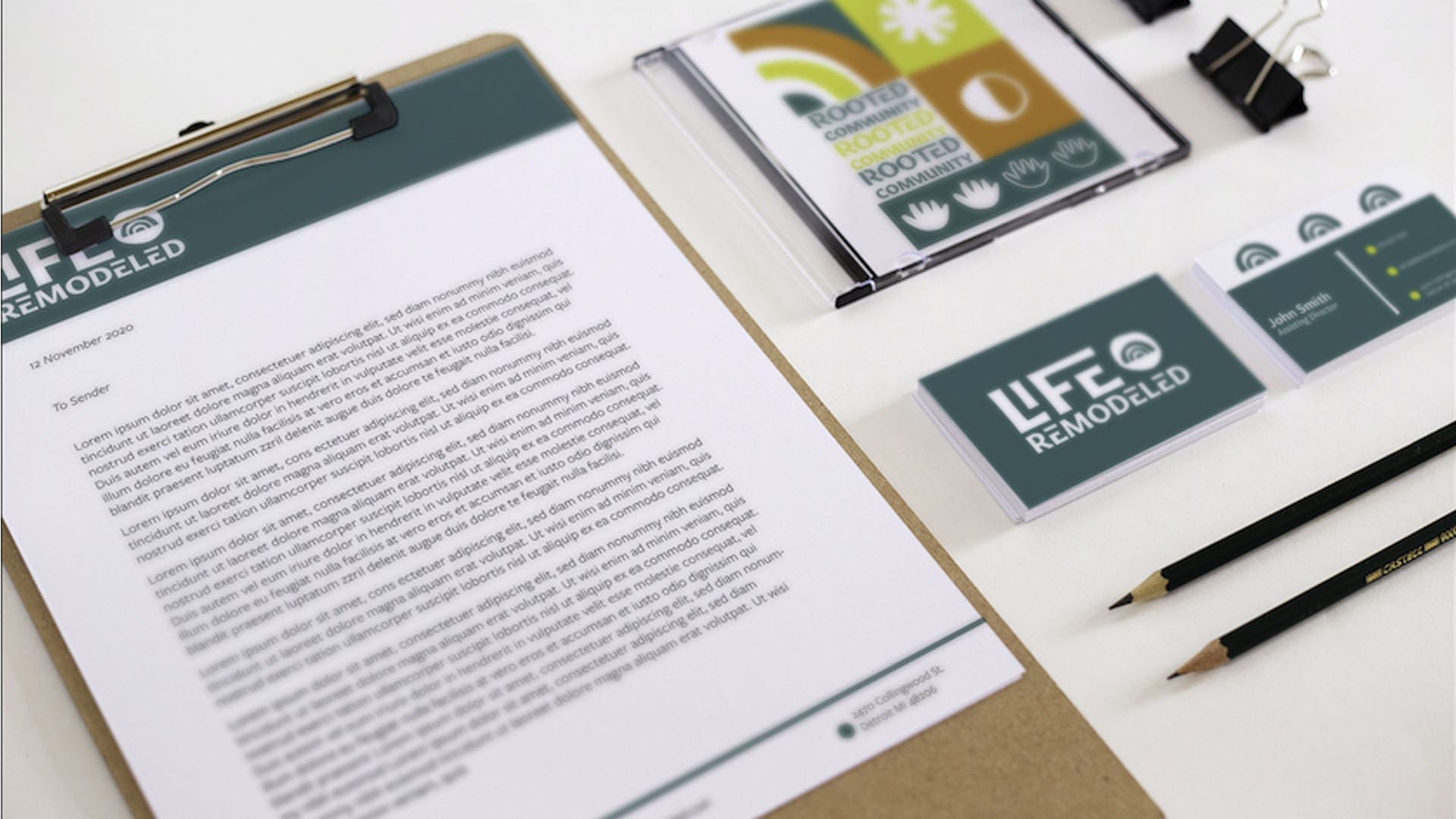 Complete rebranding of Life Remodeled. Photo showing a piece of paper with black text on a clipboard next to two black pencils, two binder clips, two dark green business cards, and a cd on a white background. Both the header of the paper and the bussiness cards show the white logo of the company with stylized text. The cd cover shows different different symbols associated with Life Remodeled, with a color scheme of yellow, white, dark green, orange, and white.