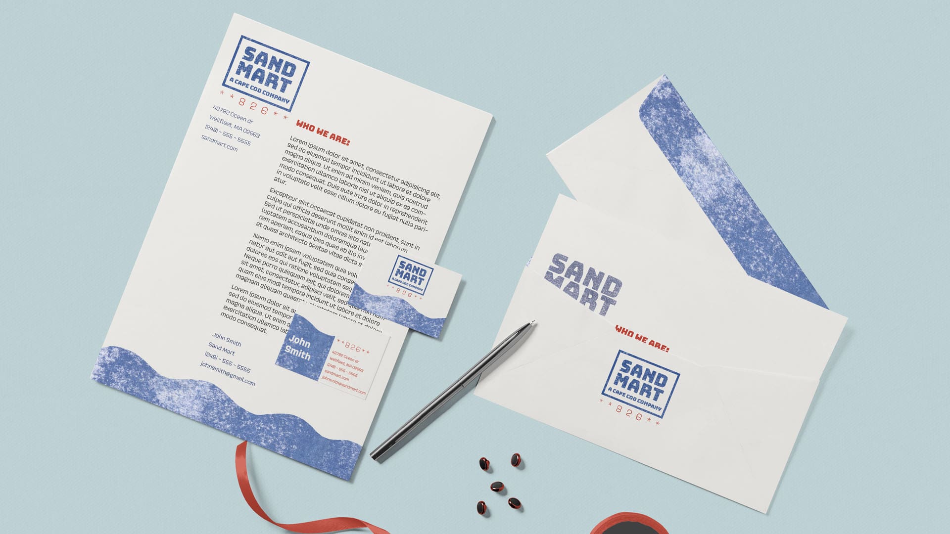 Branding for the company Sand Mart. The photo depicts a paper, bussiness card, black pen, and envelope against a pale blue background. The paper, card and envelope are white with small black text, a red header, and the blue logo of the company.