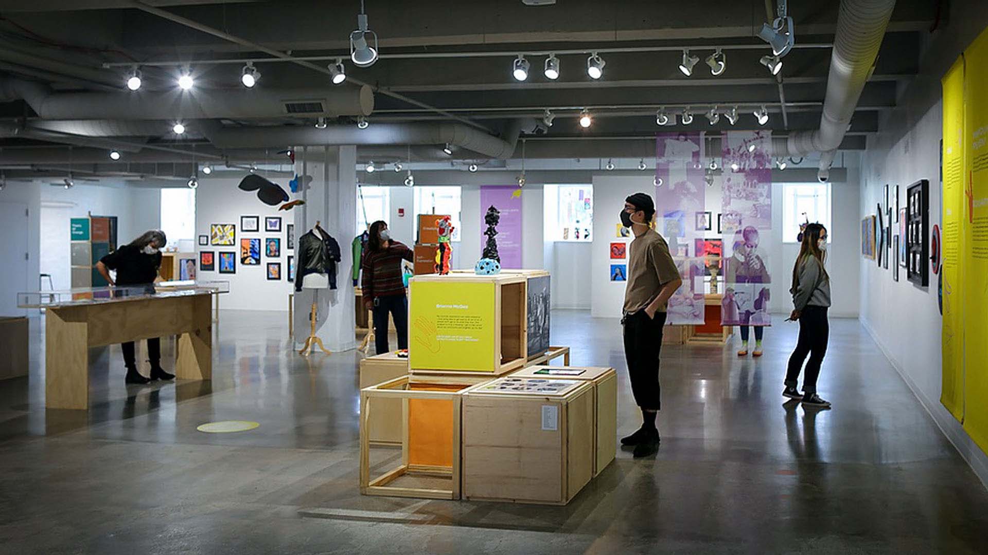 Exhibit designed to celebrate the Community Arts Partnerships 20 anniversary. Photo shows exhibit with several people milling around and looking at various art and information installations, including stacked colored boxes and purple posters.