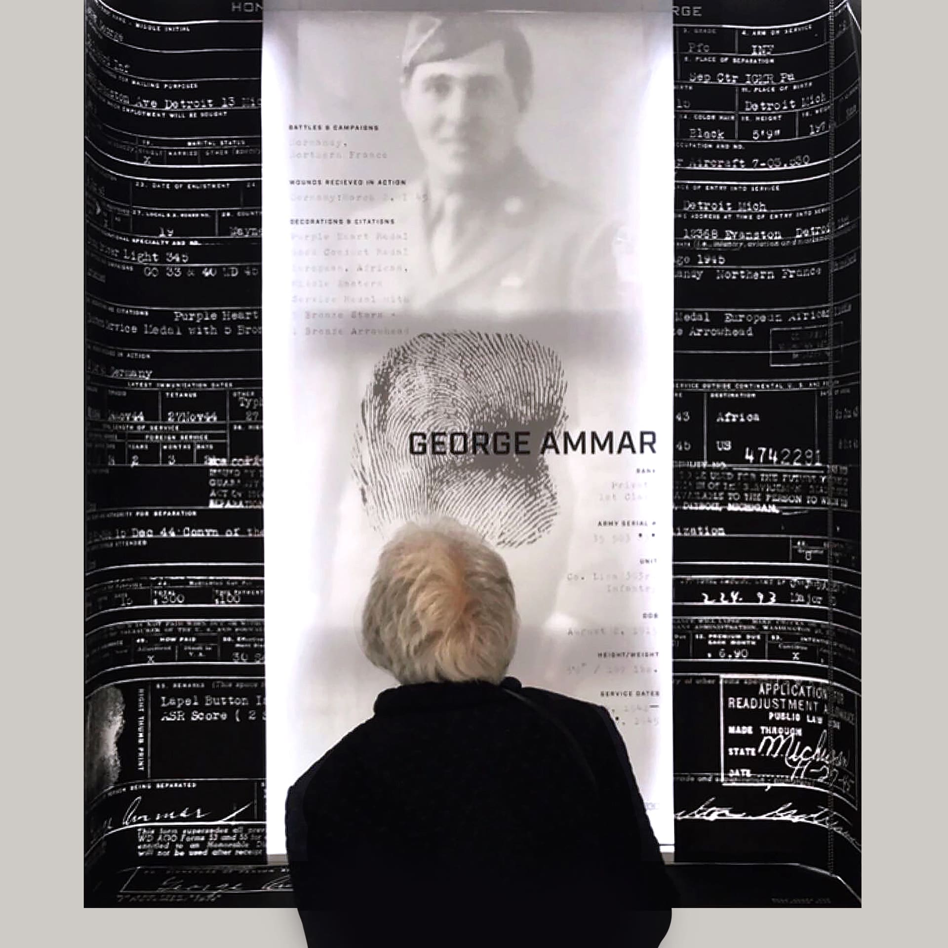 Photo of an interactive experience that shows a person looking at a large poster on a white wall. The center of the poster shows a faded black and white photo of a soldier behind a black fingerprint and black text that says "George Ammar". To the left and right are black panels with lots of white text.