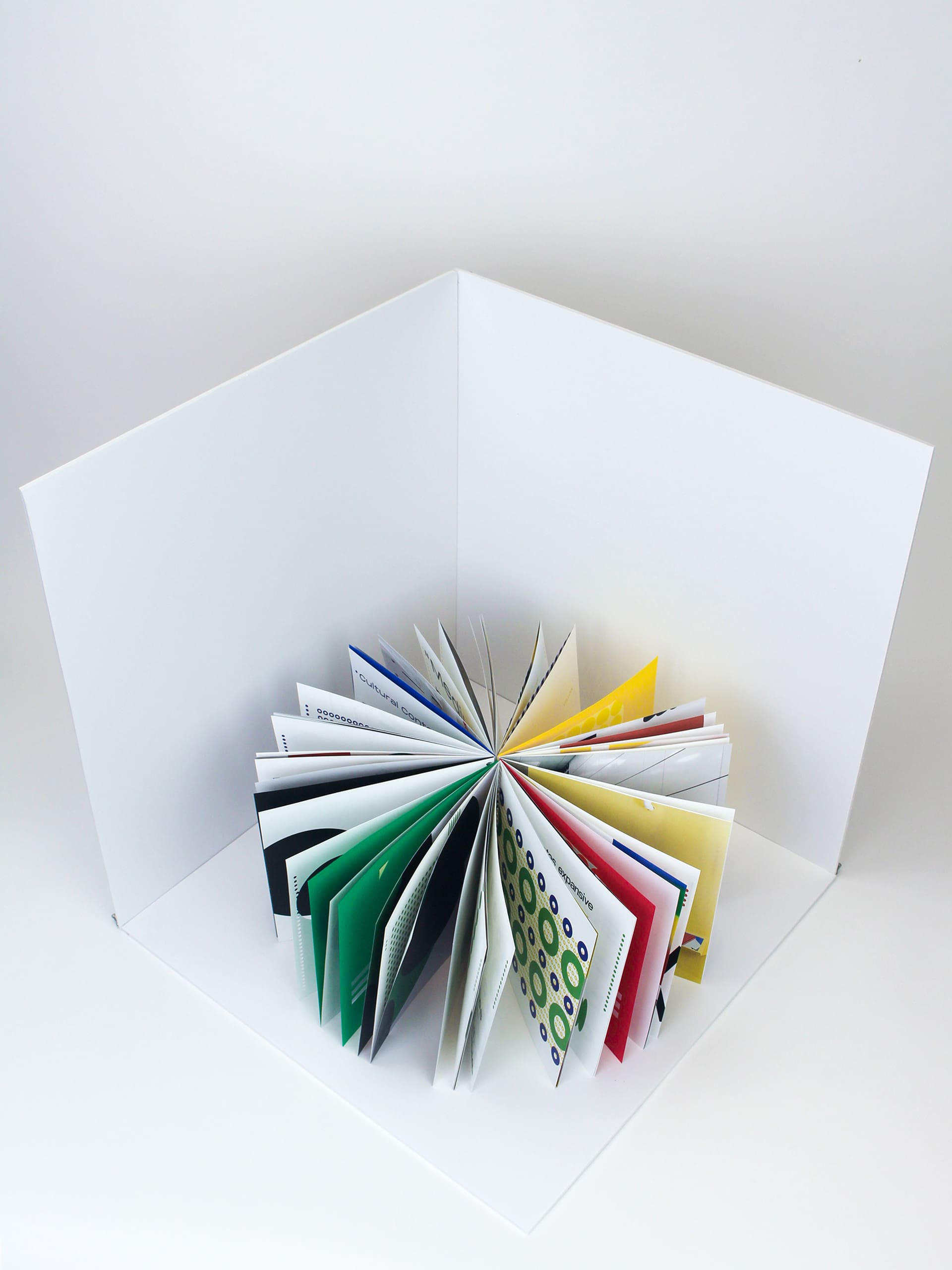 Photo of a book with yellow, red, blue, and green pages propped up and splayed out in a circle. Pictured in a white corner.