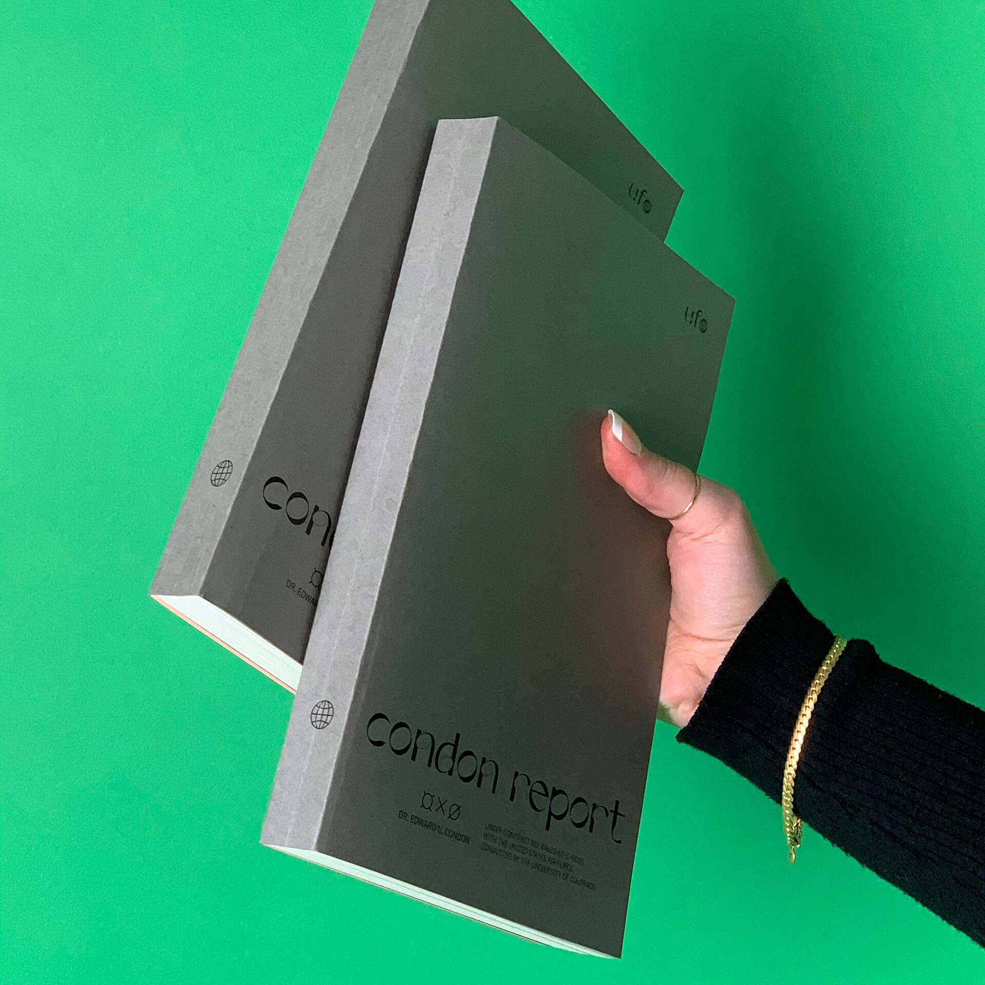 Photo of someone holding up two black books against a green wall with one hand. At the bottom of the books as the title "Condon Report" written in black glossy text.