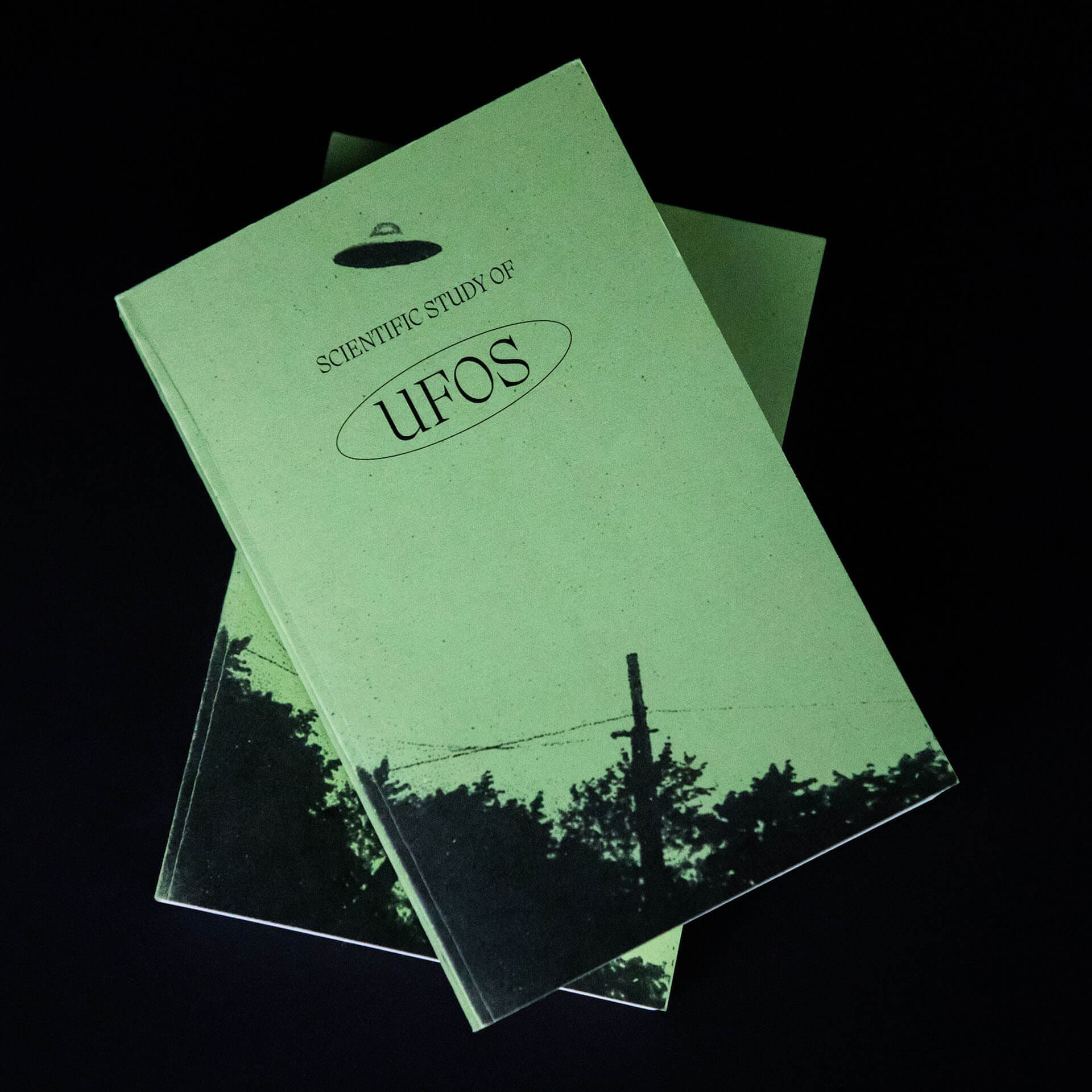Photo of two green books stacked on top of each other on a black background. The top of the book has a black title that says "Scientific Study of UFOs" below a black UFO symbol. At the bottom of the book is a black image of tree tops.