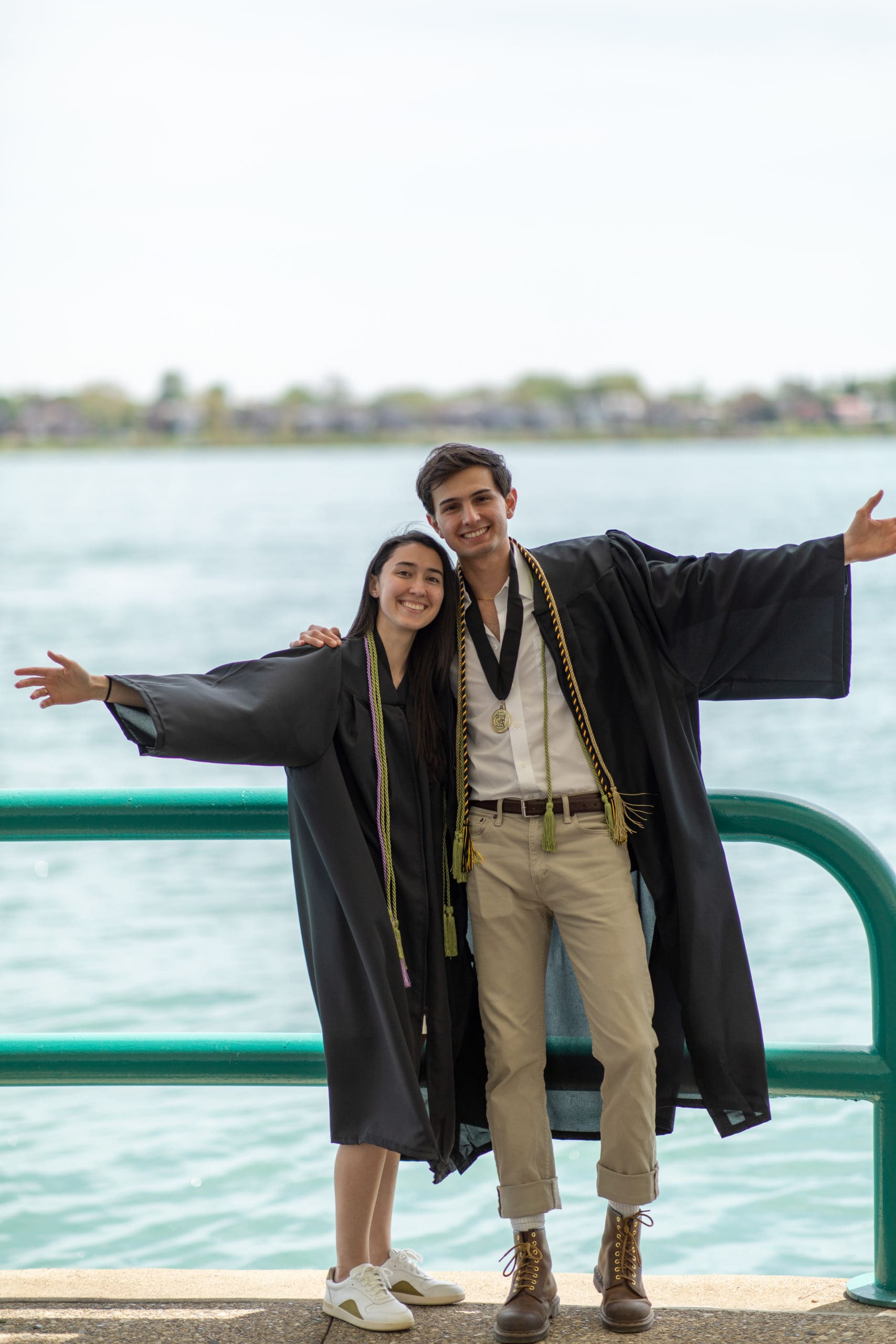 two graduating student pose with arms open in front of the Detroit River
