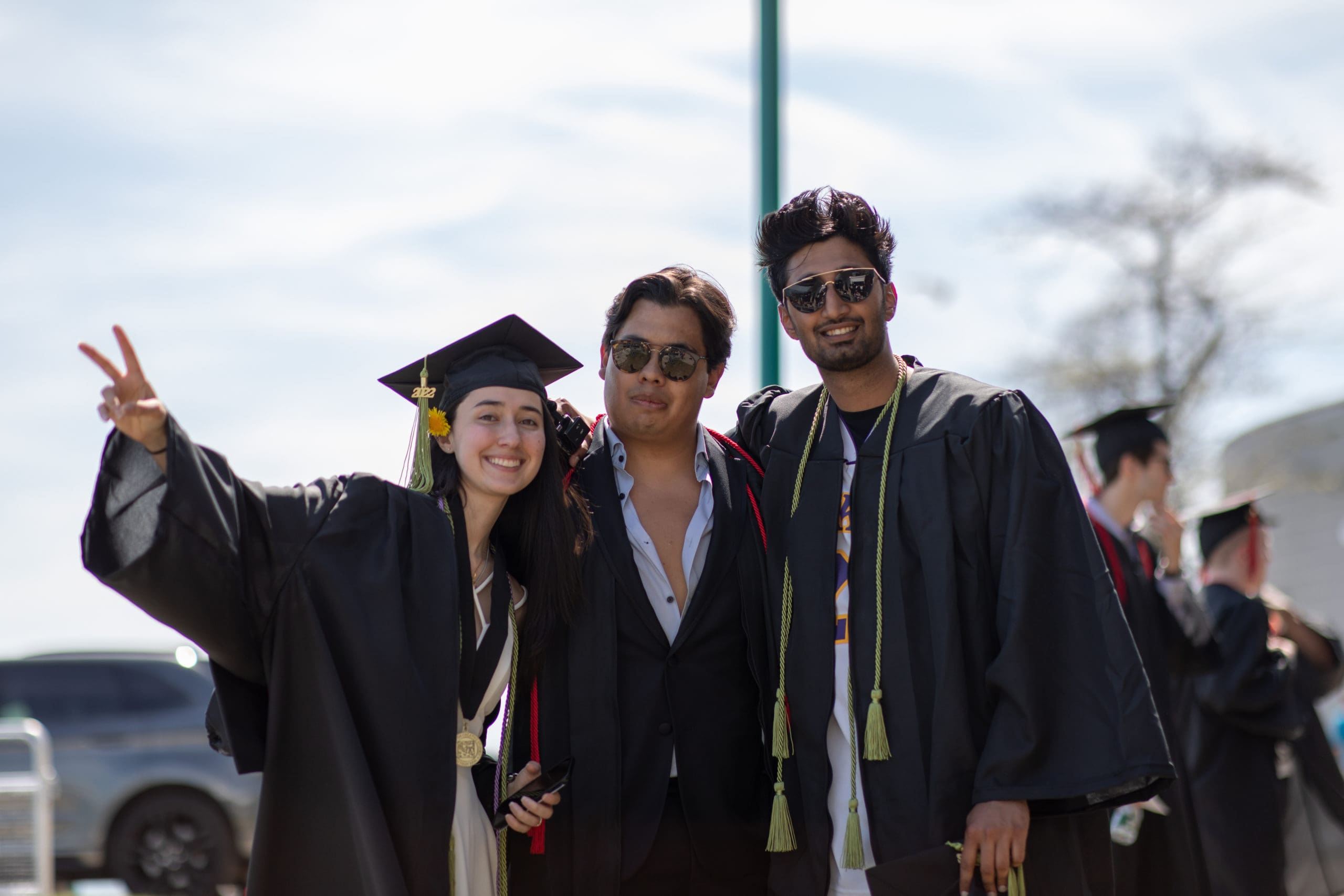Three CCS graduates in black graduation robes and multicolored cords smile and pose for a picture on a sunny day.