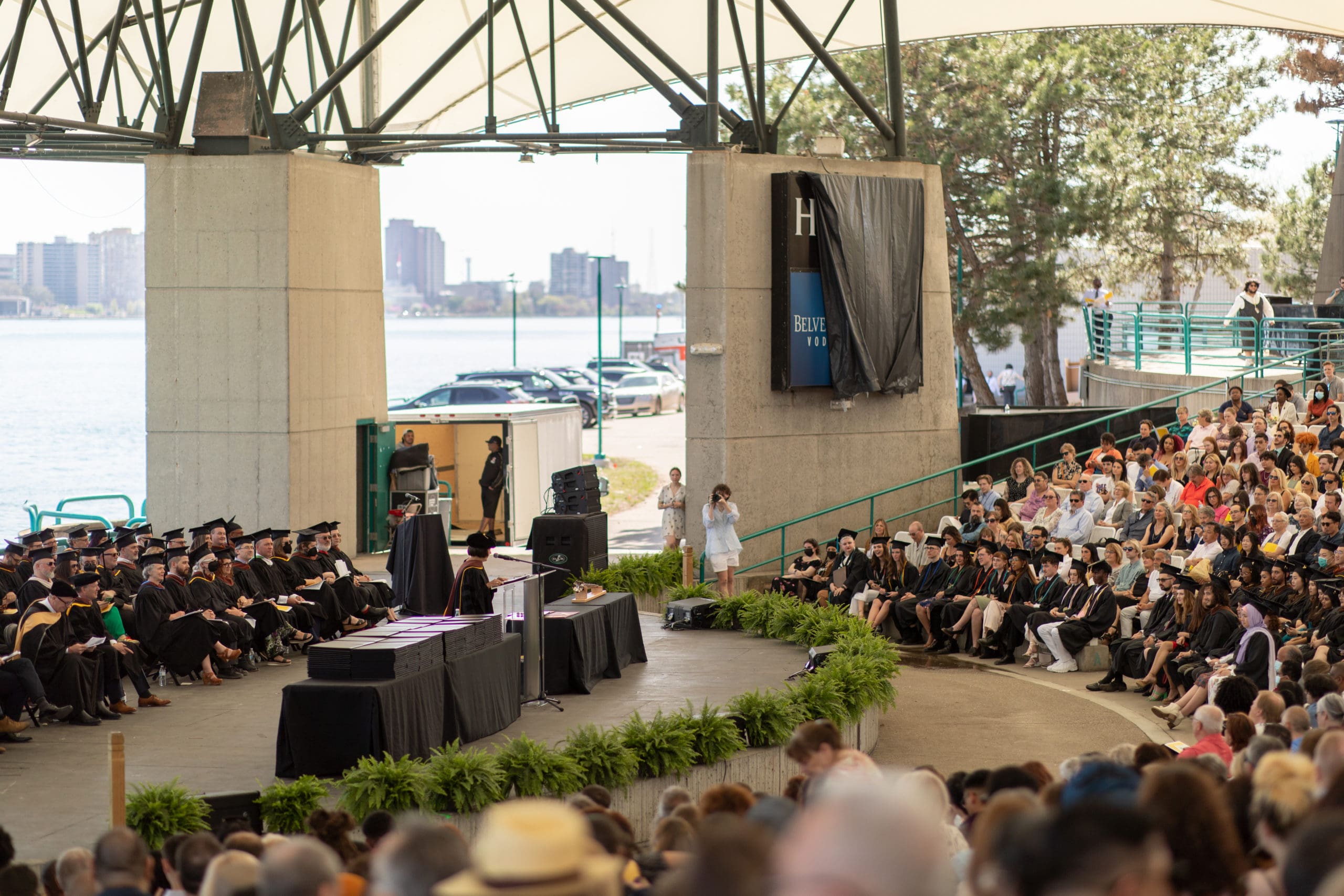 Fifth overhead view of a graduation ceremony in an outdoors theatre. A faculty member in a multicolored stole gives a speech in the center of a stage at a podium.