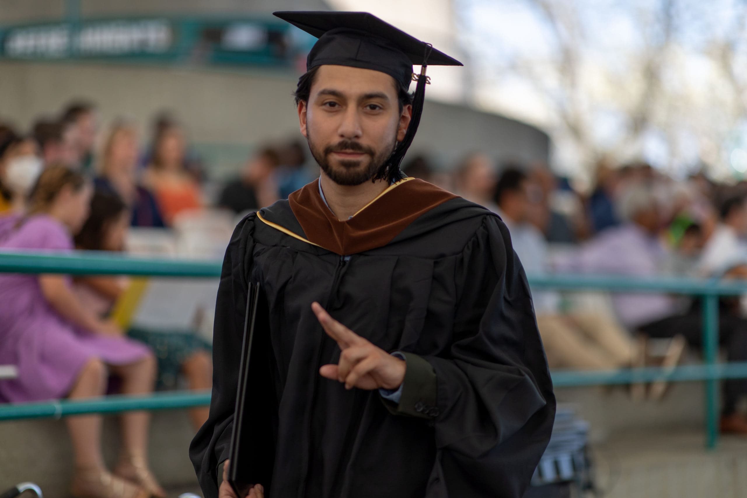Graduate student in black robes throws a peace sign to the camera while walking past the audience with their new diploma.