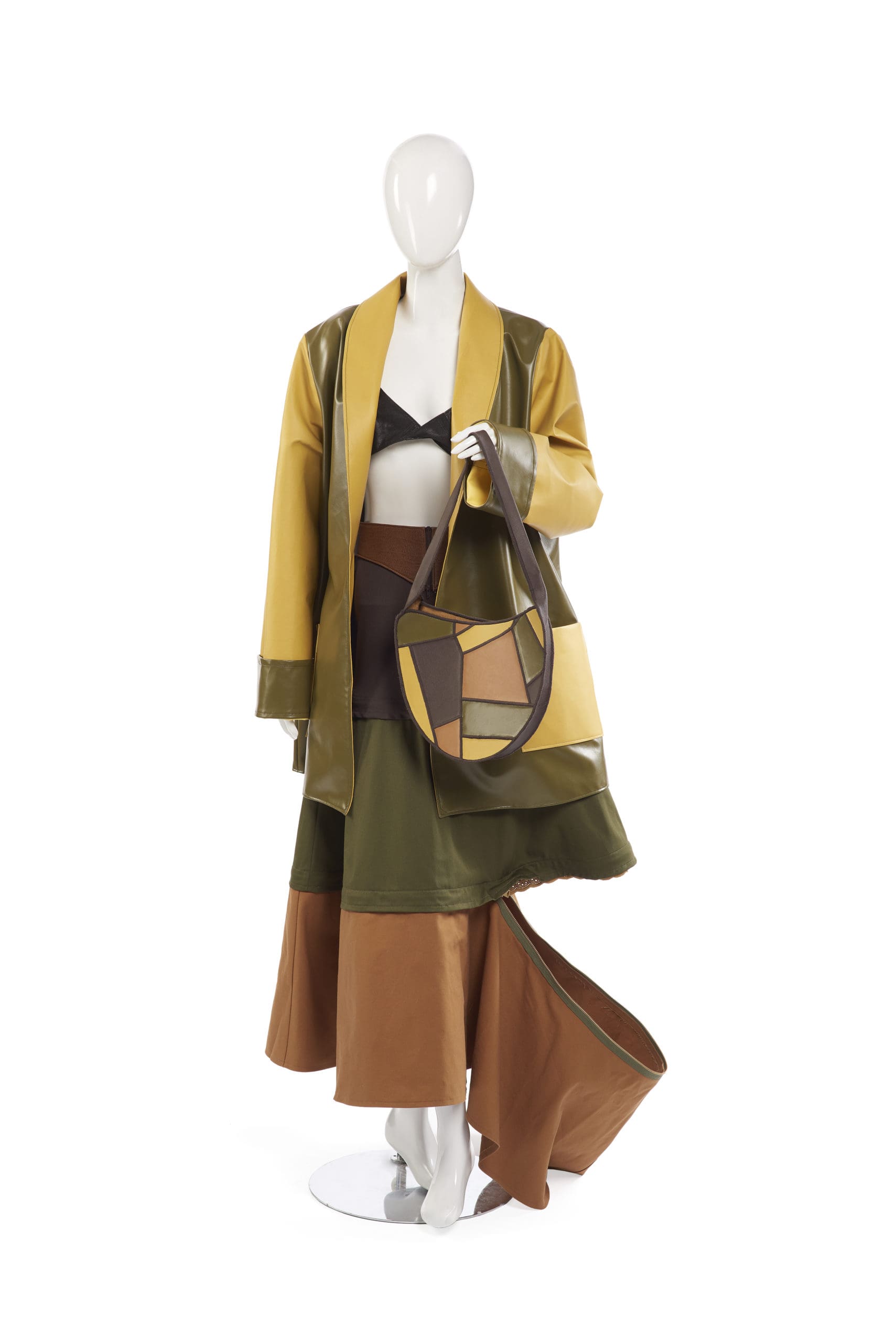 Margaret Henige. White mannequin wearing a long yellow and brown leather coat, a long skirt with separate green, brown, burnt orange and light brown layers, and holding a purse with a geometric shape design with the same colors as the dress.
