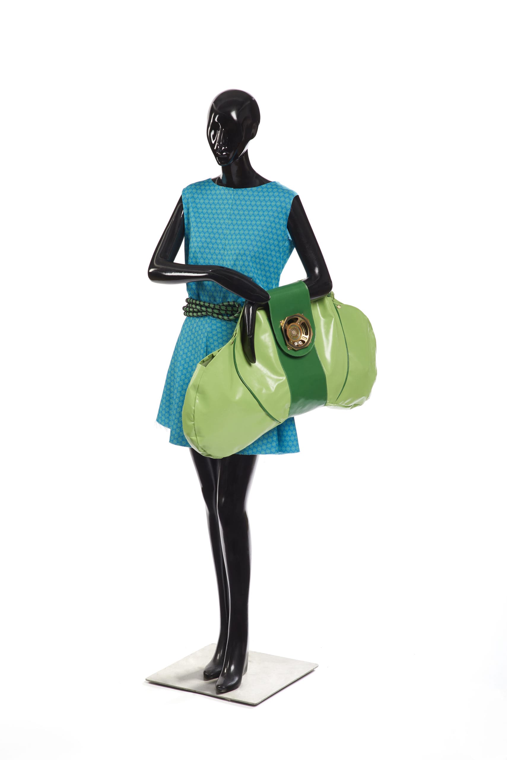 Black mannequin wearing a bright blue polka dot mini dress with a green rope belt. Also holding a large faux plastic light green bag with a wide dark green strap and a large gold button clasp
