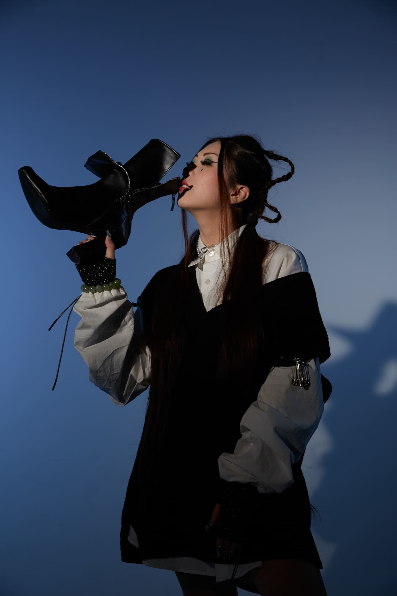 Nikki Park. Photo of a model on a blue background holding a black leather Ankle Boot with Double Tongue. They are wearing an oversized white shirt with black vest.