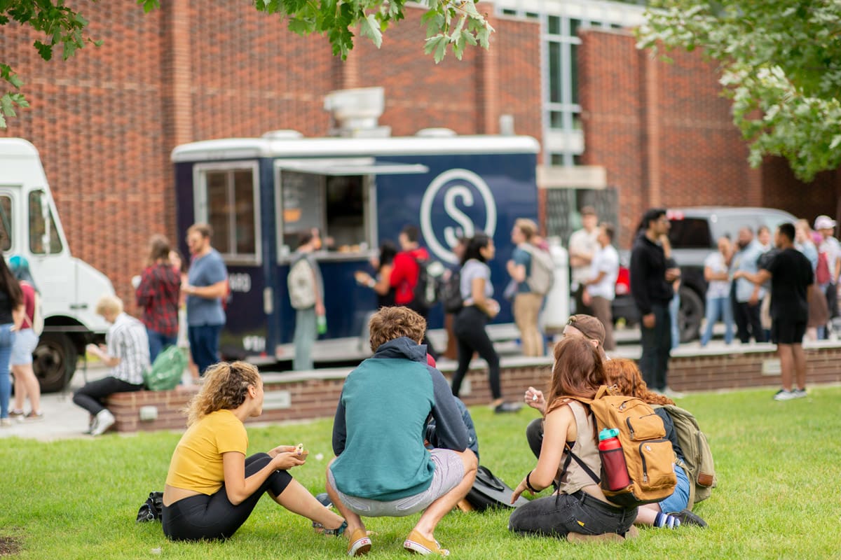 Group of students sitting together on the grass in front of the Yamasaki Building, where there is a line of food trucks offering food to the public
