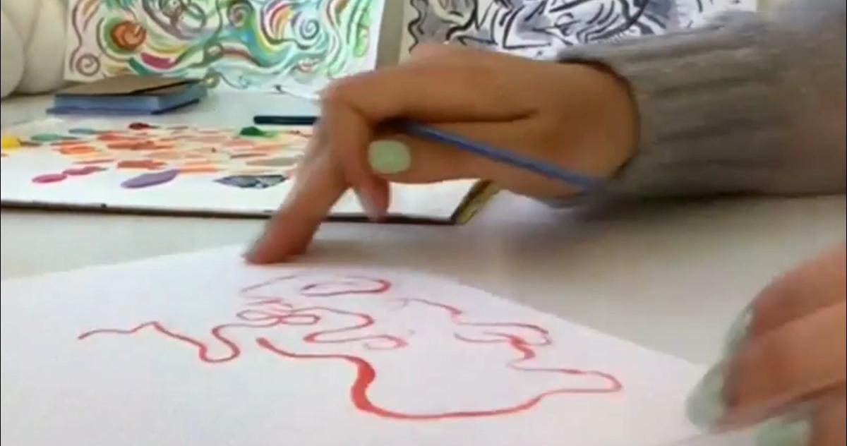 Still of a video depicting a women's hand holding a blue paintbrush over a white piece of paper with some red paint squiggles.